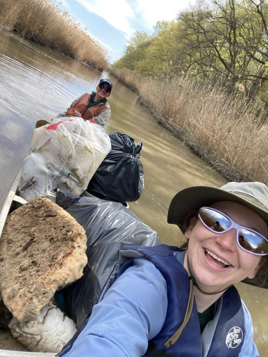 Two people paddle a canoe filled with trash bags and debris through a marsh.