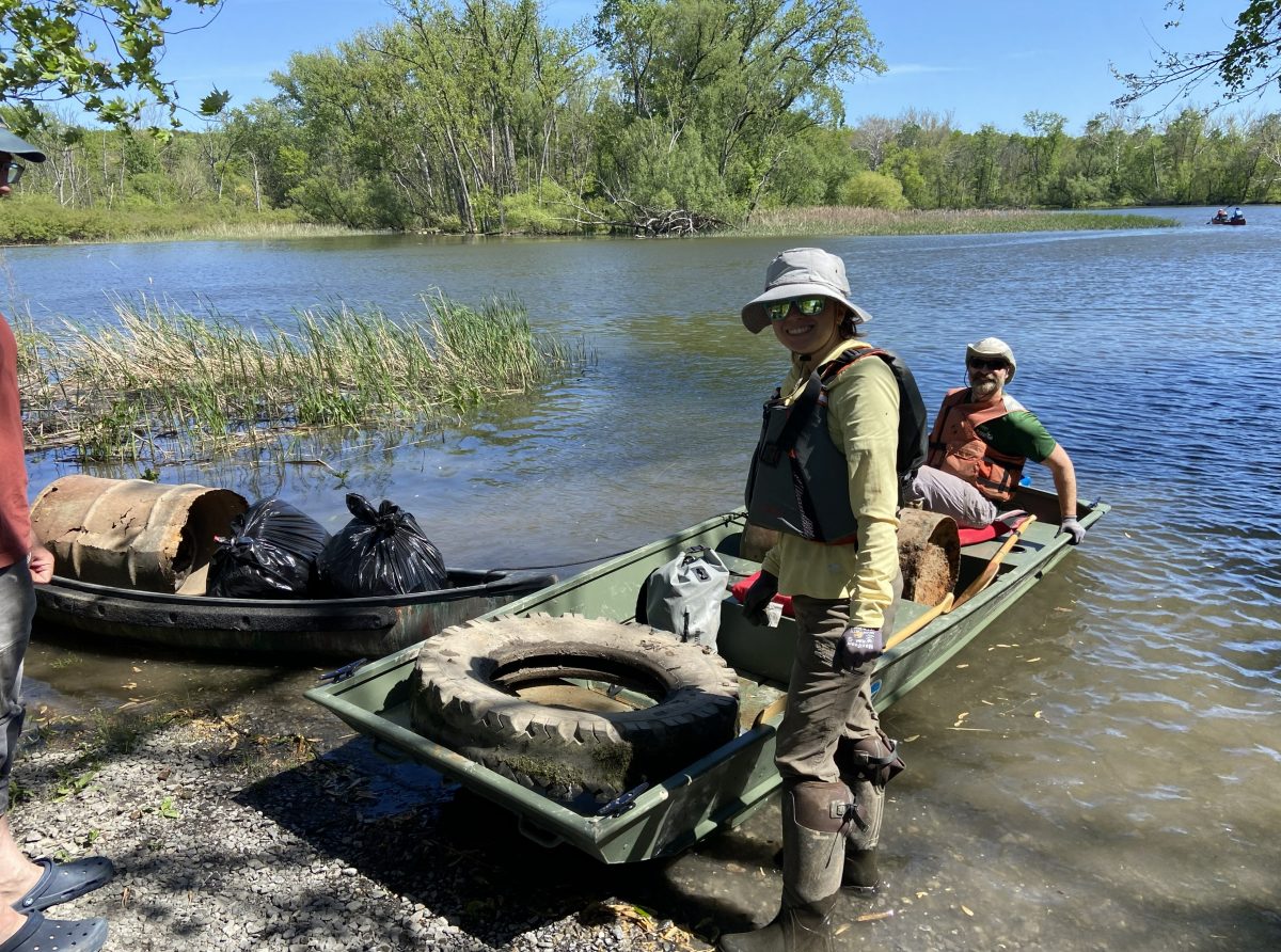 One person stands at the water's edge next to a boat. Inside the boat, one person sits next to a tire and other debris.