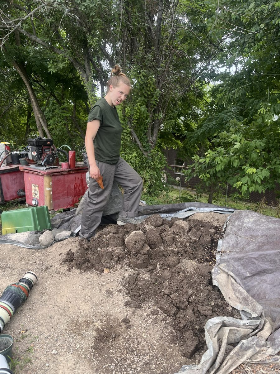 A young person stands on a large pile of soil with plastic plant pots at the bottom of the soil pile.