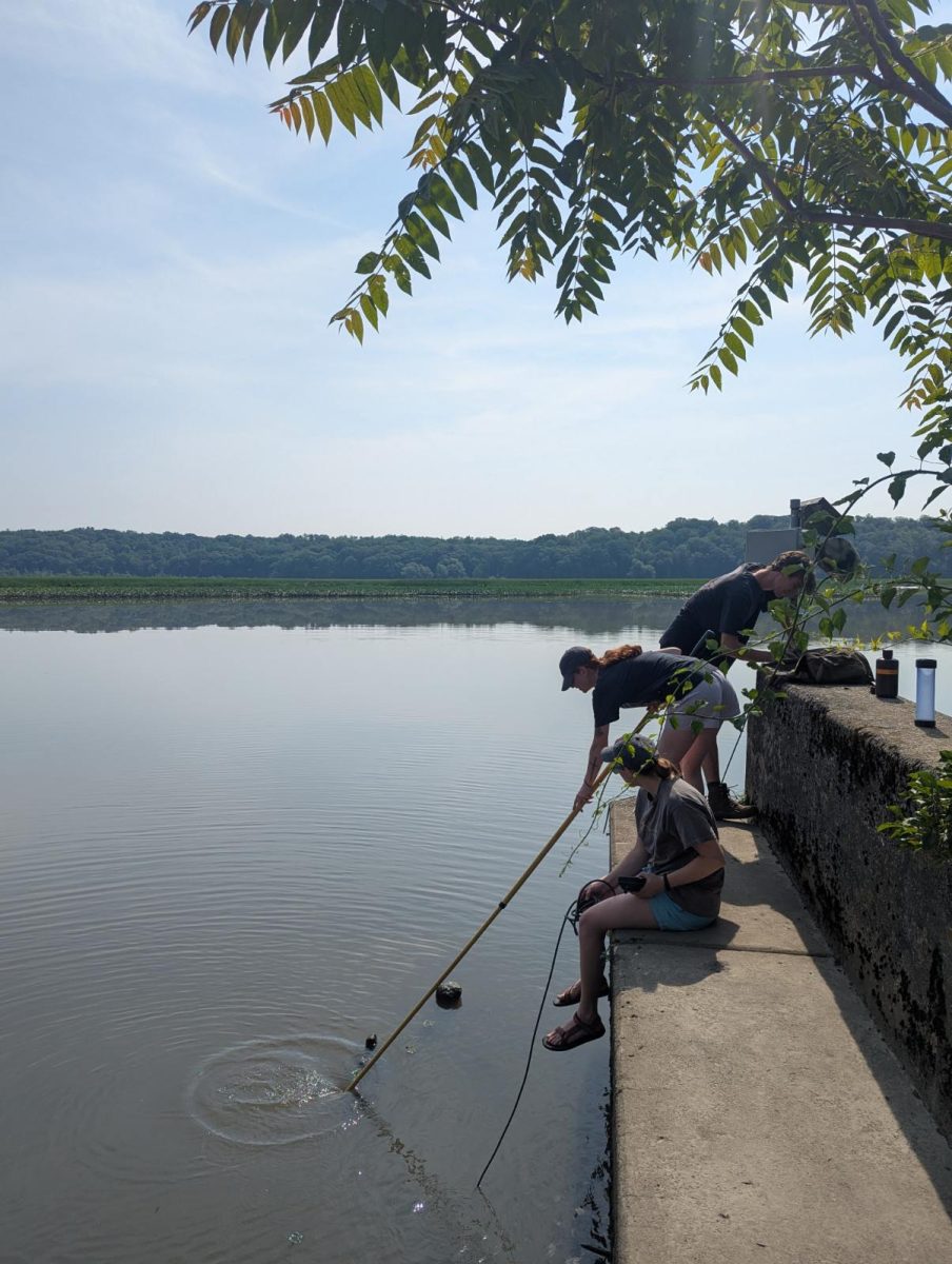 Three people work on a concrete ledge that overhangs a small bay at the edge of a tidal wetland. One person uses a long pole to collect a water sample from the bay.