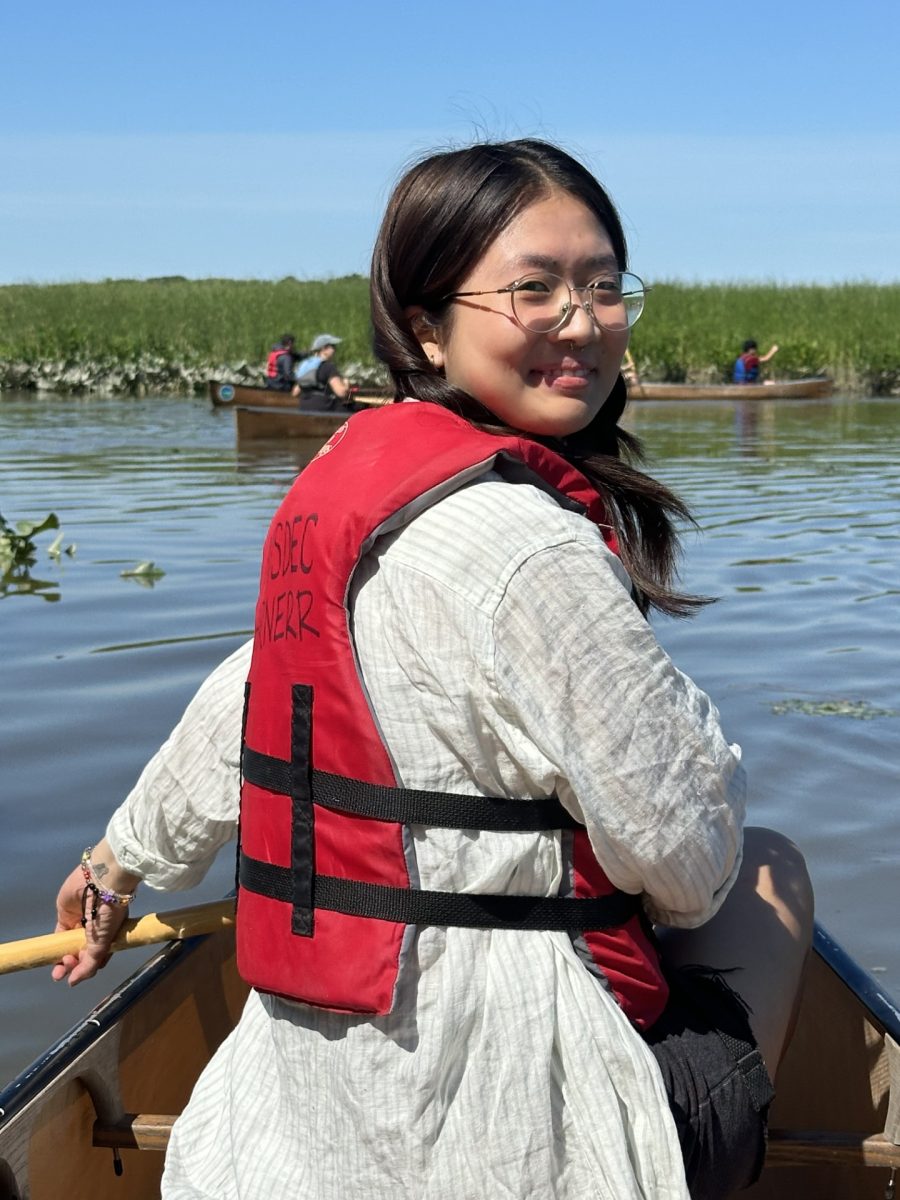 A young woman wearing a life jacket sits in a canoe and looks over her shoulder. In the background, other canoes with paddlers and marsh vegetation are visible.